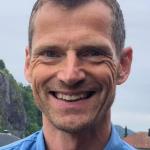 Portrett av en smilende Robert Rognli i blå skjorte utendørs med lettskyet himmel