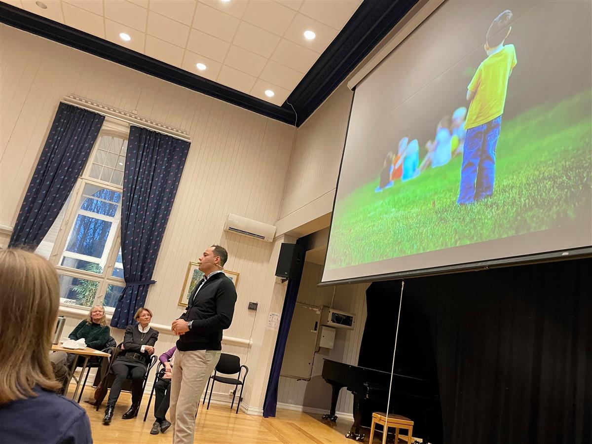 Aiman Shaqura holder foredrag for publikum, og på presentasjonen ser vi en gutt som er utenfor en gruppe barn som sitter på gresset - Klikk for stort bilde